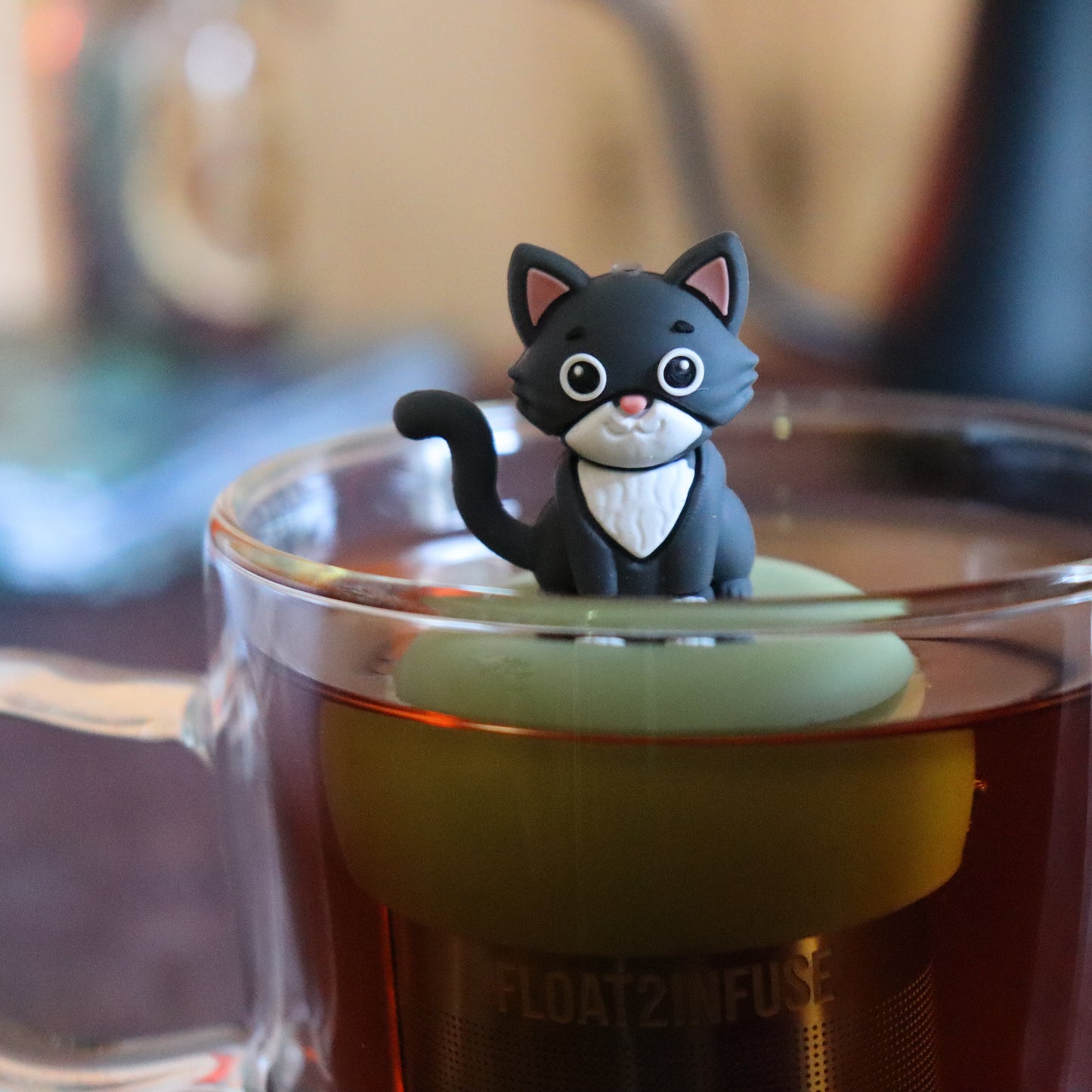 grey and white cat animal tea infuser for loose leaf tea on a green base with a fine mesh stainless steel tea strainer that reads FLOAT2INFUSE floating in a glass tea mug with tea kettle in the background