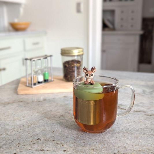 happy brown and beige deer tea infuser for loose leaf tea with a fine mesh stainless steel tea strainer that reads FLOAT2INFUSE on a green base floating in a glass tea mug
with loose leaf tea in the background