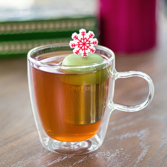 red and white holiday snowflake tea infuser for loose leaf tea with a fine mesh stainless steel tea strainer that reads FLOAT2INFUSE on a green base floating in a glass tea mug
with books in the background