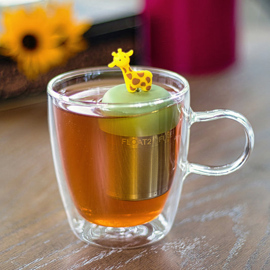yellow giraffe animal tea infuser for loose leaf tea with a fine mesh stainless steel tea strainer that reads FLOAT2INFUSE on a green base floating in a glass tea mug
with flowers in the background