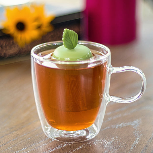 green herb leaf tea infuser for loose leaf tea with a fine mesh stainless steel tea strainer that reads FLOAT2INFUSE on a green base floating in a glass tea mug with flowers in the background