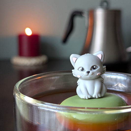 3d grey cat tea infuser for loose leaf tea with a fine mesh stainless steel tea strainer that reads FLOAT2INFUSE on a green base with a lit candle and a tea kettle in the background