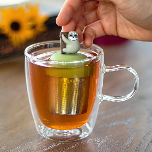 grey sloth animal climbing a tree tea infuser for loose leaf tea with a fine mesh stainless steel strainer that reads FLOAT2INFUSE on a green base floating in a glass tea mug with flowers in the background