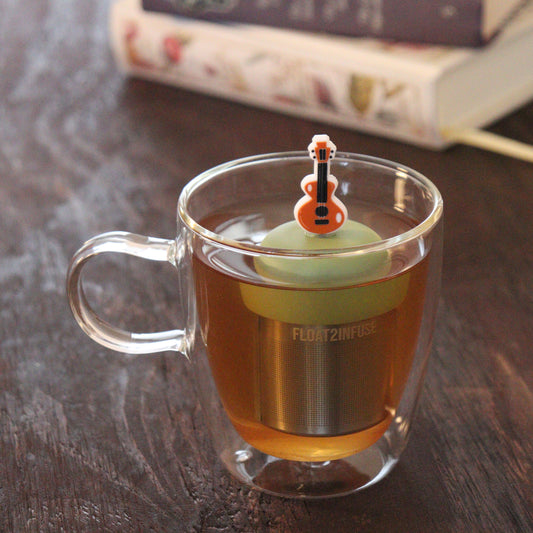 brown guitar tea infuser for loose leaf tea with a fine mesh stainless steel tea infuser that reads FLOAT2INFUSE on a green base floating in a glass tea mug with books in the background