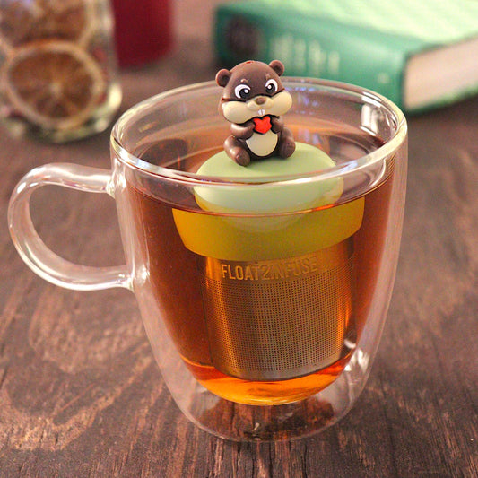 beaver animal holding a red heart tea infuser for loose leaf tea with a fine mesh stainless steel tea infuser that reads FLOAT2INFUSE on a green base floating in a glass tea mug with loose leaf tea and books in the background