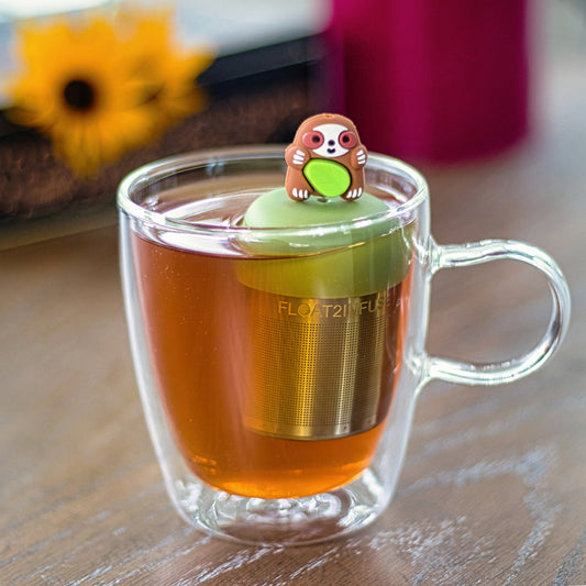 brown sloth animal holding a green leaf infuser for loose leaf tea with a fine mesh stainless steel tea strainer that reads FLOAT2INFUSE on a green base floating in a glass tea mug
with flowers in the background