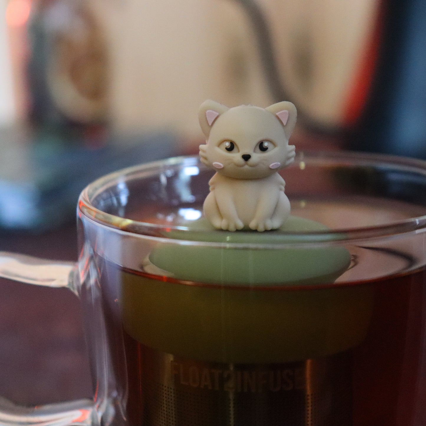 ivory cat animal tea infuser for loose leaf tea on a green base with a fine mesh stainless steel tea strainer that reads FLOAT2INFUSE floating in a glass tea mug with tea kettle in the background