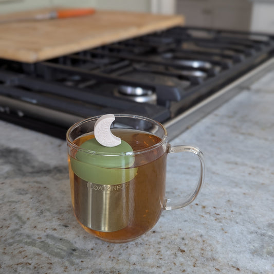 ivory crescent moon with raised floral pattern tea infuser for loose leaf tea with fine mesh stainless steel tea strainer that reads FLOAT2INFUSE on a green base floating in a glass tea mug with a stove in the background
