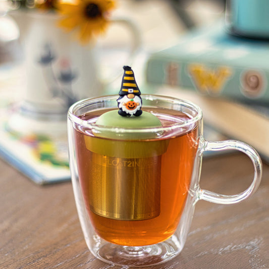 gnome with long white beard wearing black with yellow stripes and holding a jack-o-lantern pumpkin tea infuser for loose leaf tea with a fine mesh stainless steel tea strainer that reads FLOAT2INFUSE on a green base floating in a glass tea mug
with books in the background