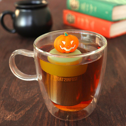 Jack-o-lantern pumpkin tea infuser for loose leaf tea with a fine mesh stainless steel tea strainer that reads FLOAT2INFUSE on a green base floating in a glass tea mug with a cauldron and books in the background