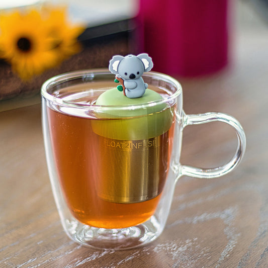 grey koala animal on a tree branch tea infuser for loose leaf tea with a fine mesh stainless steel tea strainer that reads FLOAT2INFUSE on a green base floating in a glass tea mug
with flowers in the background