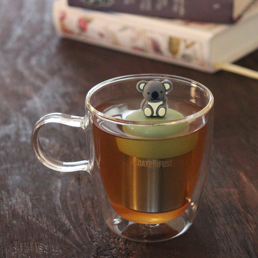 waving grey koala animal tea infuser for loose leaf tea with a fine mesh stainless steel tea infuser that reads FLOAT2INFUSE on a green base floating in a glass tea mug with books in the background