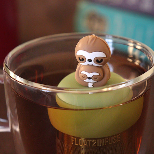 Mom and baby sloth tea infuser for loose leaf tea with a fine mesh stainless steel tea strainer
that reads FLOAT2INFUSE on a green base floating in a glass tea mug
with a tea canister and books in the background