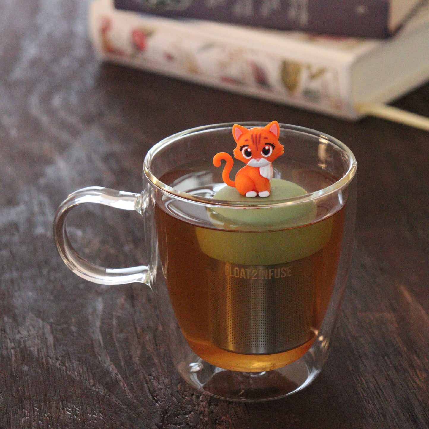 orange cat animal tea infuser for loose leaf tea with a fine mesh stainless steel tea infuser that reads FLOAT2INFUSE on a green base floating in a glass tea mug with books in the background