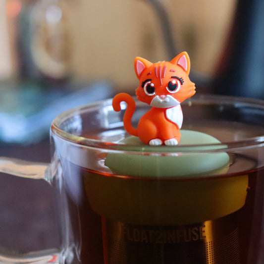 orange cat animal tea infuser for loose leaf tea on a green base with a fine mesh stainless steel tea strainer that reads FLOAT2INFUSE floating in a glass tea mug with tea kettle in the background