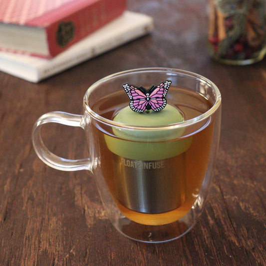 pink animal tea infuser for loose leaf tea with a fine mesh stainless steel tea infuser that reads FLOAT2INFUSE on a green base floating in a glass tea mug with books and loose leaf tea in the background