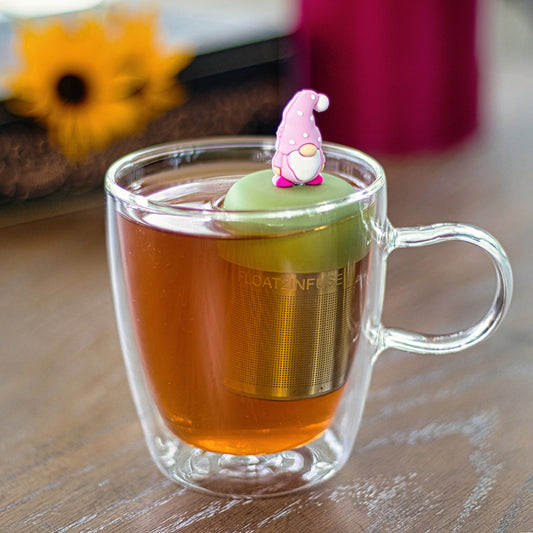 gnome with a long white beard and wearing light light pink clothing with white polka dots on the hat tea infuser for loose leaf tea with a fine mesh stainless steel strainer that reads FLOAT2INFUSE on a green base floating in a glass tea mug
with a flowers in the background