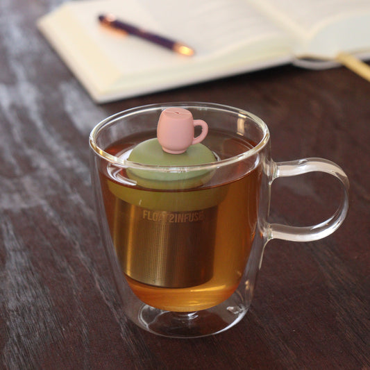 pink tea cup tea infuser for loose leaf tea with a fine mesh stainless steel tea infuser that reads FLOAT2INFUSE on a green base floating in a glass tea mug with notebook and pen in the background