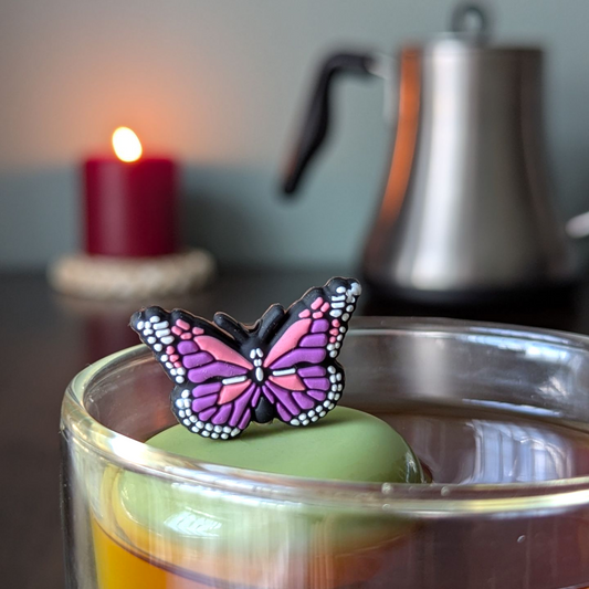 Purple and pink butterfly tea infuser for loose leaf tea with a fine mesh stainless steel tea strainer that reads FLOAT2INFUSE on a green base with a lit candle and a tea kettle in the background