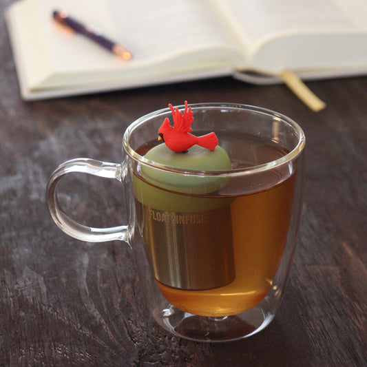red cardinal bird animal tea infuser for loose leaf tea with a fine mesh stainless steel tea infuser that reads FLOAT2INFUSE on a green base floating in a glass tea mug with notebook and pen in the background