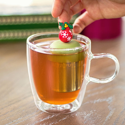 red ball christmas ornament with white snowflakes and green holly plant tea infuser for loose leaf tea with a fine mesh stainless steel tea strainer that reads FLOAT2INFUSE on a green base floating in a glass tea mug with books in the background