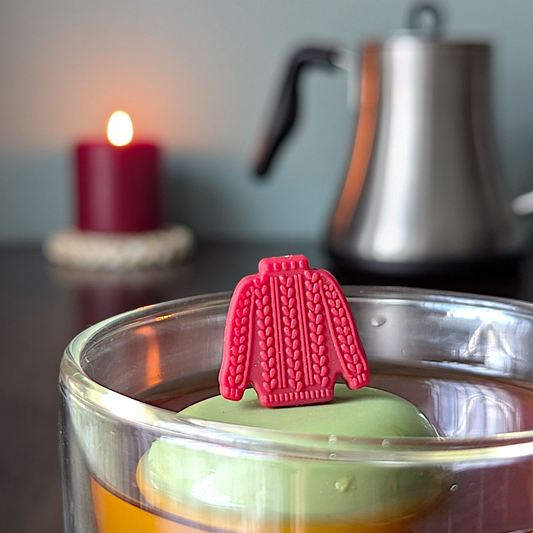 red knit sweater tea infuser for loose leaf tea with a fine mesh stainless steel tea strainer that reads FLOAT2INFUSE on a green base with a lit candle and a tea kettle in the background