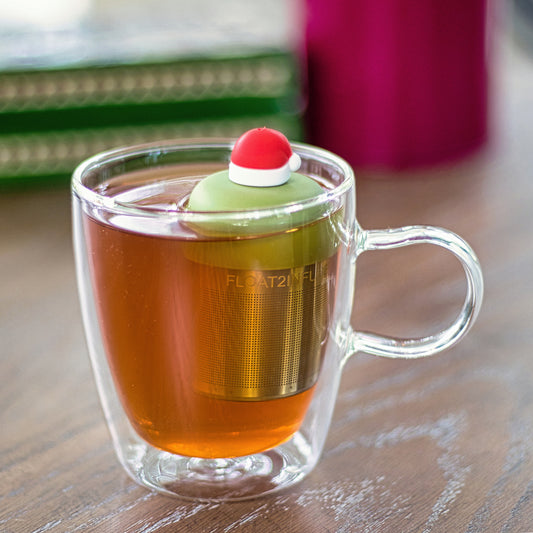 red santa hat with white trim tea infuser for loose leaf tea with a fine mesh stainless steel tea strainer
that reads FLOAT2INFUSE on a green base floating in a glass tea mug
with books in the background