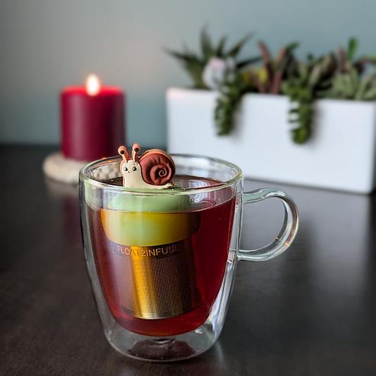 smiling brown snail tea infuser for loose leaf tea with a fine mesh stainless steel tea strainer that reads FLOAT2INFUSE on a green base with a lit candle and a plant in the background