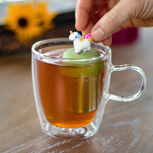 3d white unicorn with a rainbow mane and gold horn tea infuser for loose leaf tea with a fine mesh stainless steel strainer that reads FLOAT2INFUSE on a green base floating in a glass tea mug with flowers in the background