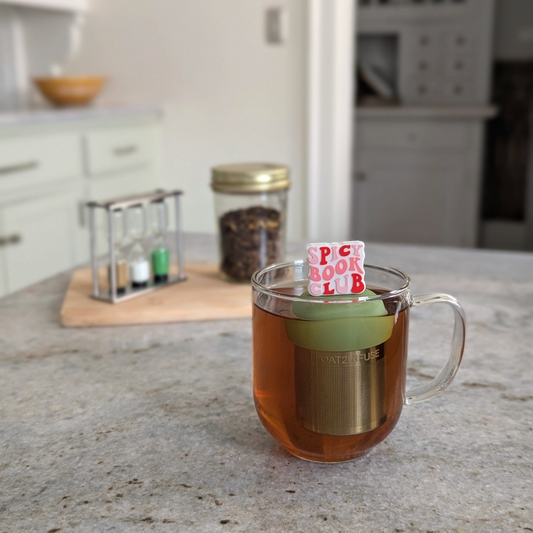 white square with text "spicy book club" in pink and red letters tea infuser for loose leaf tea with a fine mesh stainless steel strainer that reads FLOAT2INFUSE on a green base floating in a glass tea mug with loose leaf tea in the background