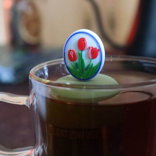 red and pink tulips framed in blue oval tea infuser for loose leaf tea with a fine mesh stainless steel tea strainer that reads FLOAT2INFUSE on a green base floating in a glass tea mug with tea kettle in the background