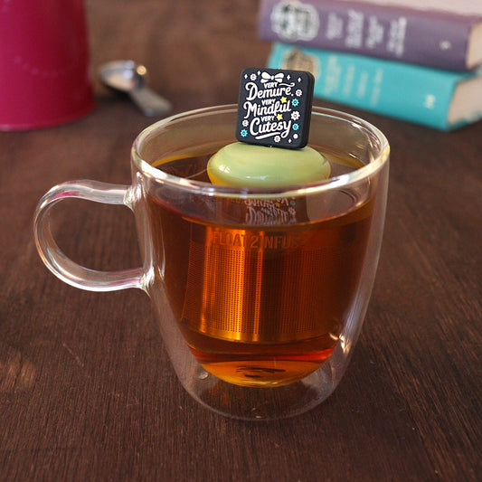 Black square with pink text that reads "very demure, very mindful, very cutesty" with pink and teal flowers and a pink bow tea infuser for loose leaf tea with a fine mesh stainless steel tea strainer that reads FLOAT2INFUSE on a green base with a tea canister and books in the background
