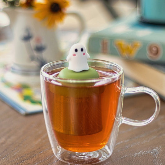 white ghost with cute pink cheeks tea infuser for loose leaf tea with a fine mesh stainless steel tea strainer
that reads FLOAT2INFUSE on a green base floating in a glass tea mug
with books in the background