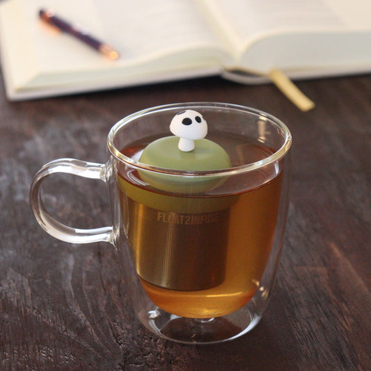white and black mushroom tea infuser for loose leaf tea with a fine mesh stainless steel tea infuser that reads FLOAT2INFUSE on a green base floating in a glass tea mug with notebook and pen in the background