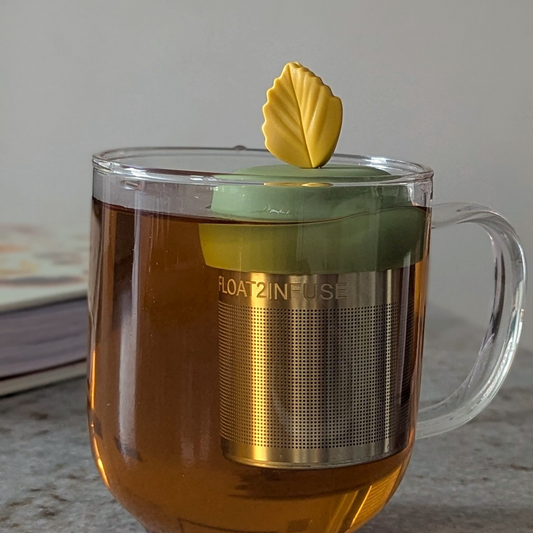 yellow fall leaf tea infuser for loose leaf tea with a fine mesh stainless steel tea strainer that reads FLOAT2INFUSE on a green base floating in a glass tea mug with a book in the background