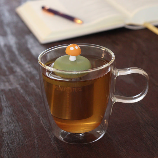 yellow mushroom tea infuser for loose leaf tea with a fine mesh stainless steel tea infuser that reads FLOAT2INFUSE on a green base floating in a glass tea mug with notebook and pen in the background