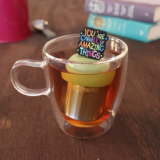 Text that reads "You are capable of amazing things" in bright rainbow font tea infuser for loose leaf tea with a fine mesh stainless steel tea strainer that reads FLOAT2INFUSE on a green base with a tea canister and books in the background