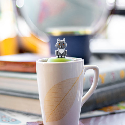 grey wolf animal tea infuser for loose leaf tea with a fine mesh stainless steel tea strainer that reads FLOAT2INFUSE on a green base floating in a white tea mug with books in the background