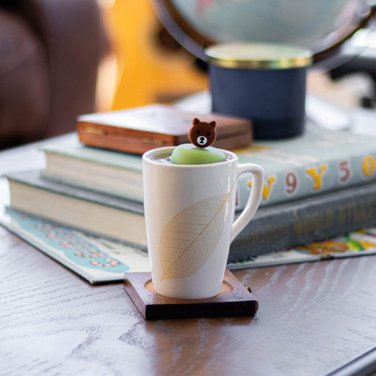 brown bear tea infuser for loose leaf tea with a fine mesh stainless steel tea strainer that reads FLOAT2INFUSE on a green base floating in a tall white tea mug on a coffee table with books in the background