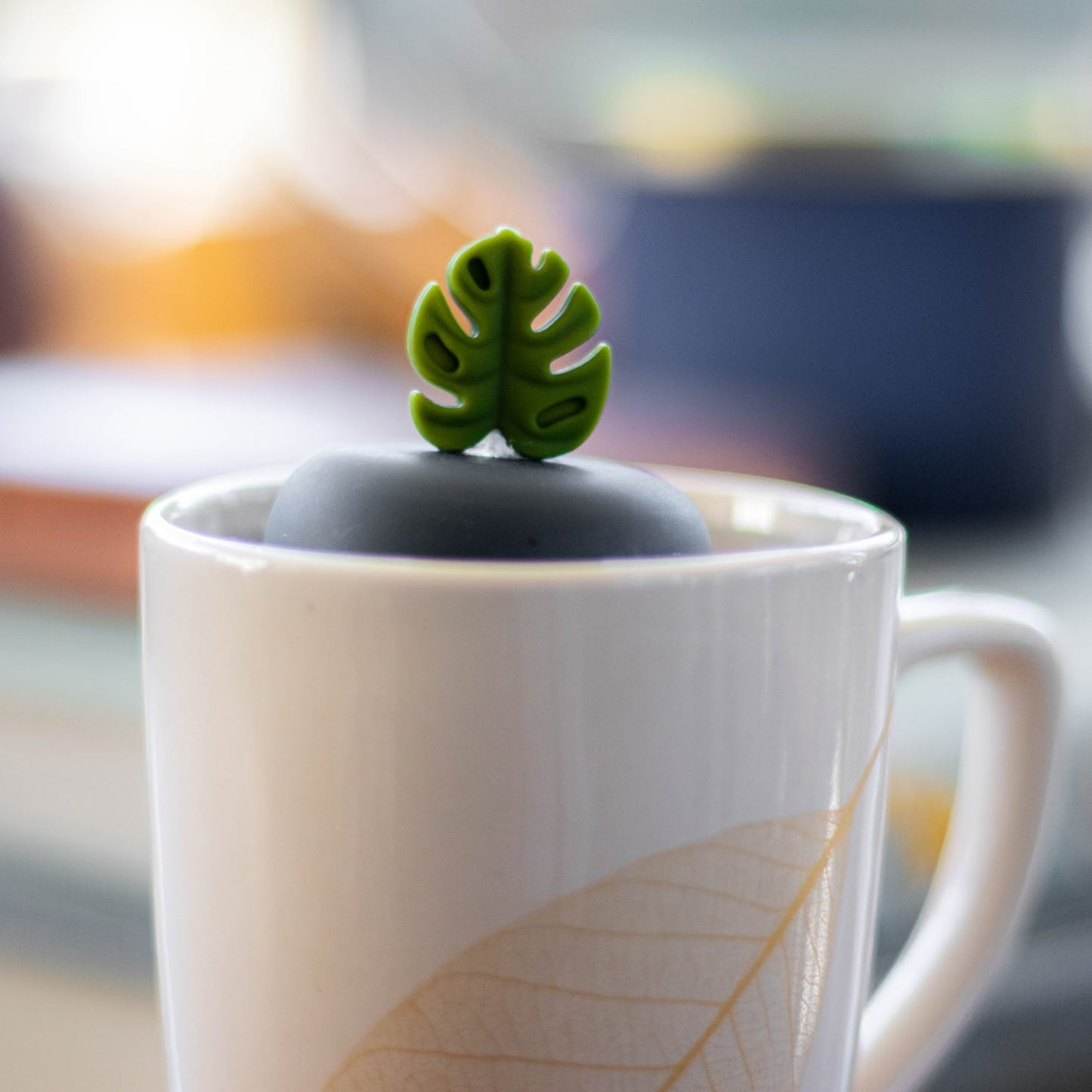 army green monstera leaf tea infuser for loose leaf tea with a fine mesh stainless steel strainer that reads FLOAT2INFUSE on a black base floating in a white tea mug with books in the background