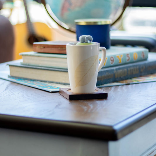 grey elephant tea infuser for loose leaf tea  with a fine mesh stainless steel strainer that reads FLOAT2INFUSE on a green base floating in a white tea cup with books, a globe, and guitar in the background