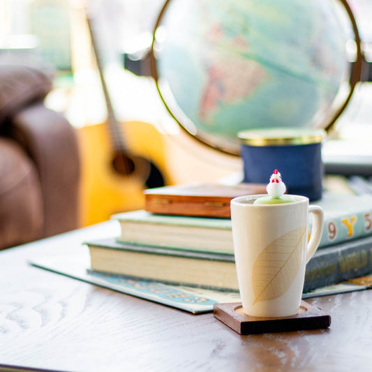 white chicken tea infuser for loose leaf tea  with a fine mesh stainless steel strainer that reads FLOAT2INFUSE on a green base floating in a white tea cup with books, a globe, and guitar in the background