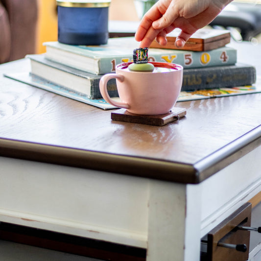 black square with shelved books, flowers, and vines and text "booktrovert" tea infuser for loose leaf tea with a fine mesh stainless steel tea strainer that reads FLOAT2INFUSE on a green base floating in a pink tea mug with books in the background
