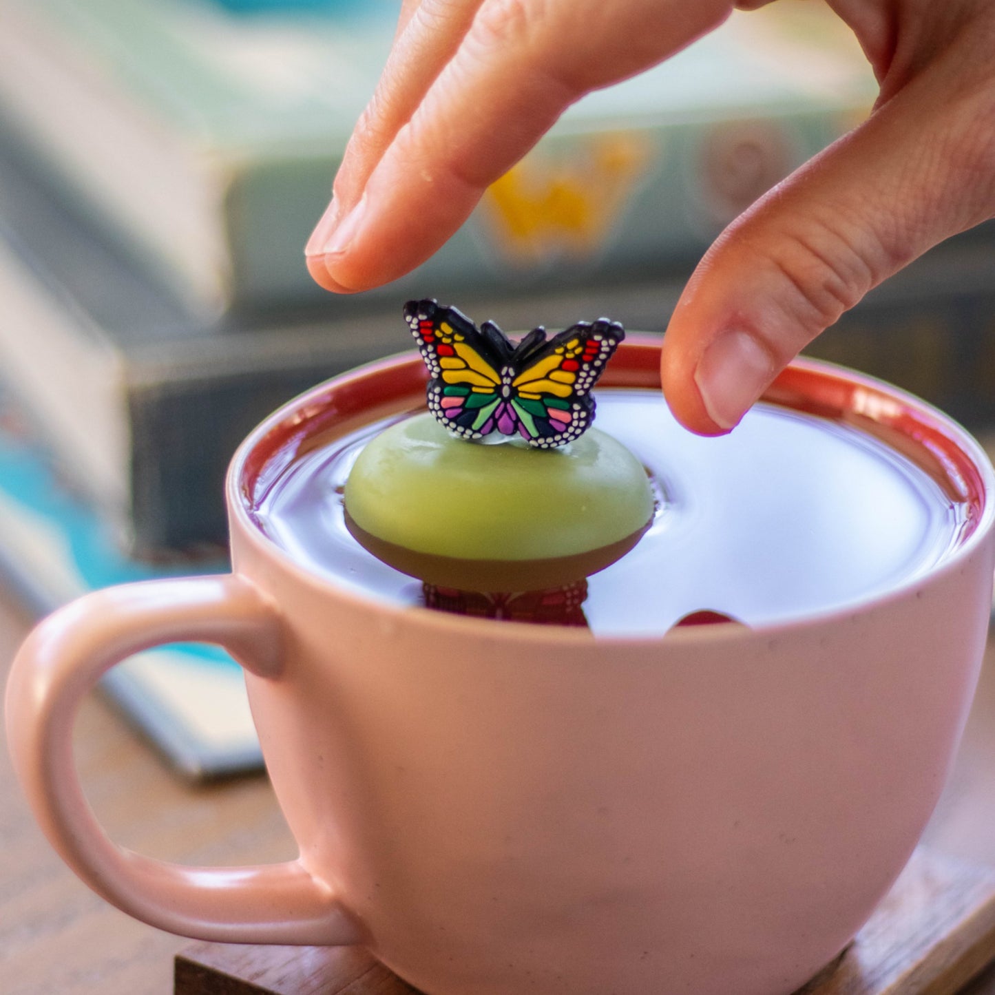 rainbow butterfly animal tea infuser for loose leaf tea with a fine mesh stainless steel tea strainer that reads FLOAT2INFUSE on a green base floating in a pink tea mug with books in the background