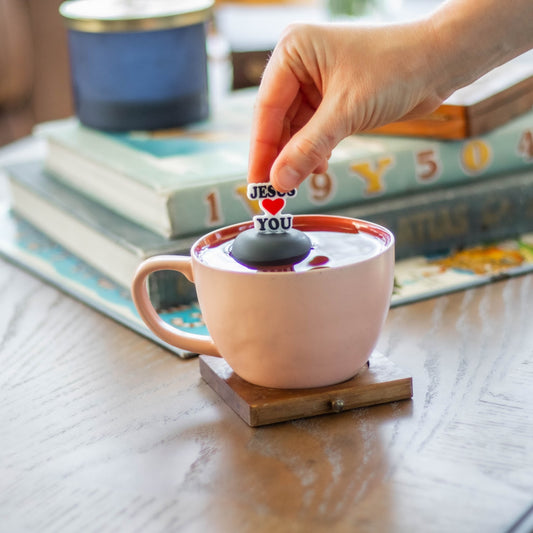 text that reads "Jesus loves you" with a heart instead of the word love tea infuser for loose leaf tea with fine mesh stainless steel tea strainer that reads FLOAT2INFUSE on a black base floating in a pink tea cup with books in the background