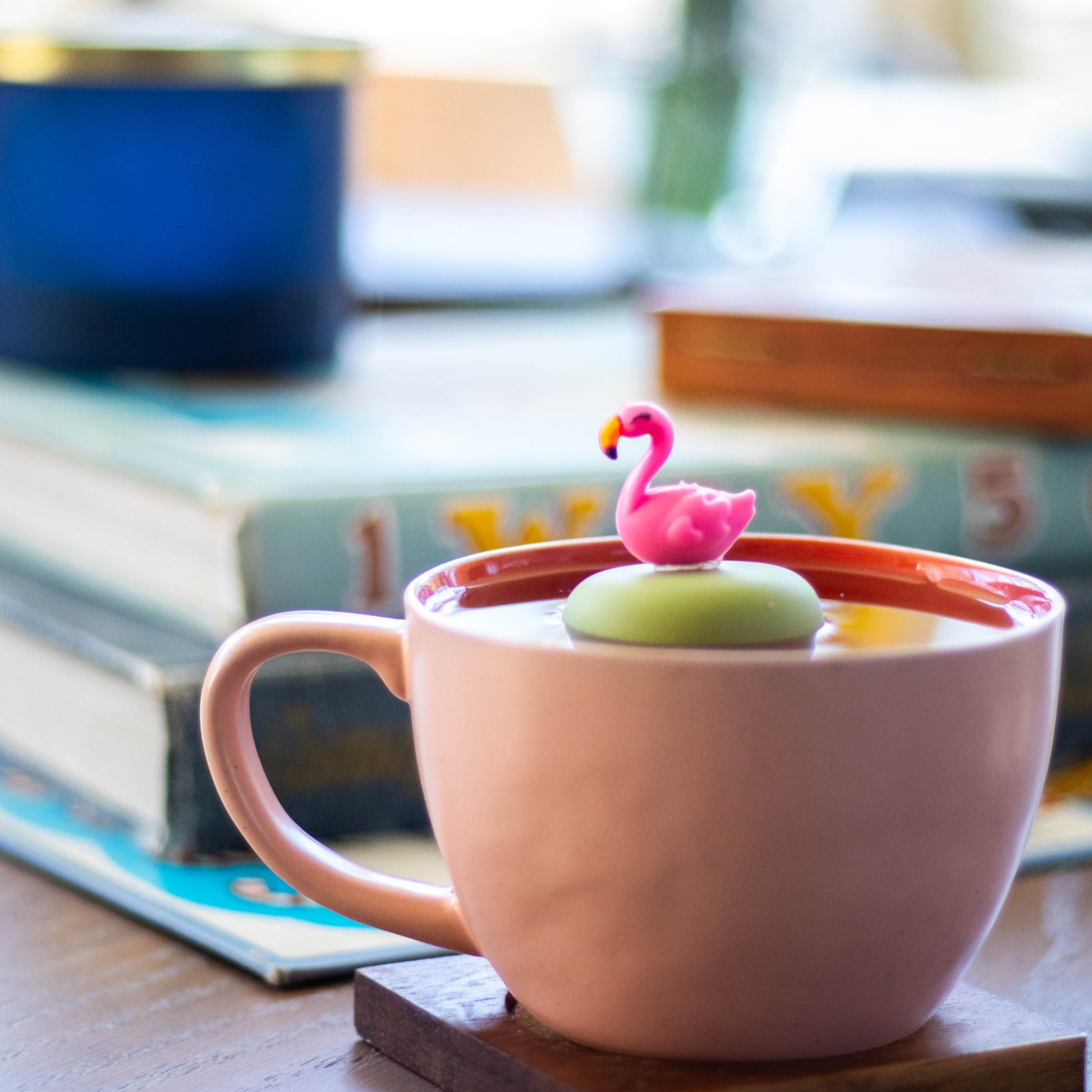 pink flamingo tea infuser for loose leaf tea  with a fine mesh stainless steel strainer that reads FLOAT2INFUSE on a green base floating in a pink tea cup with books in the background