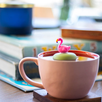 pink flamingo tea infuser for loose leaf tea  with a fine mesh stainless steel strainer that reads FLOAT2INFUSE on a green base floating in a pink tea cup with books in the background