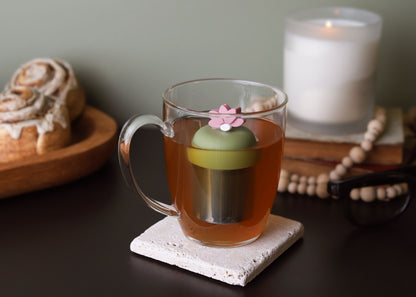 warm pink lotus flower tea infuser for loose leaf tea with a fine mesh stainless steel tea strainer that reads FLOAT2INFUSE on a green base floating in a glass tea mug with cinnamon rolls in the background