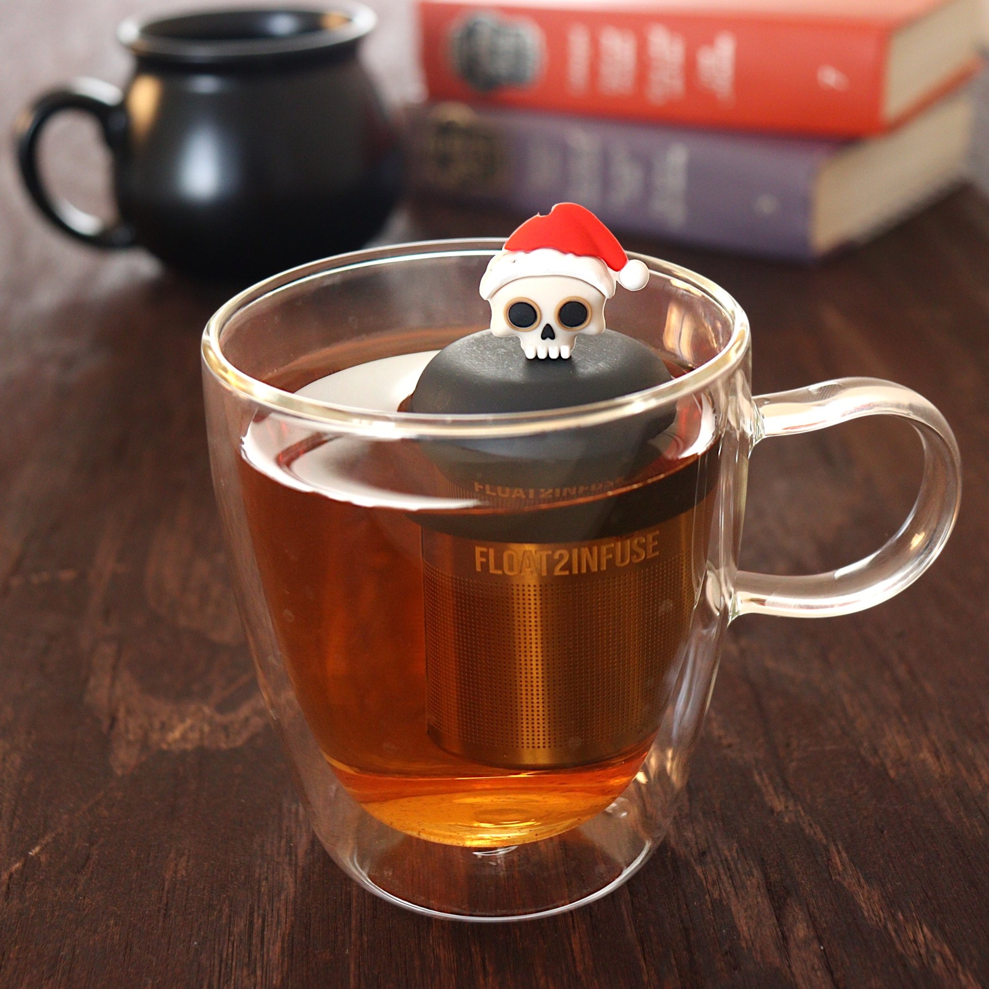 skull with a santa hat tea infuser for loose leaf tea  with a fine mesh stainless steel tea infuser that reads FLOAT2INFUSE on a black base floating in a glass tea mug with a black cauldron mug and books in the background