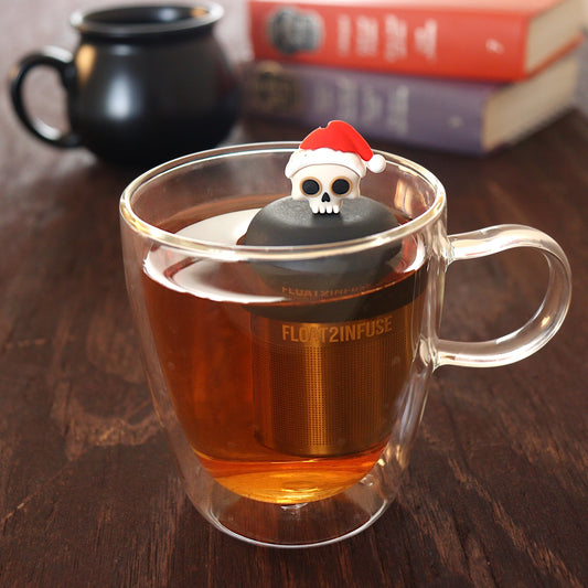 skull with a santa hat tea infuser for loose leaf tea  with a fine mesh stainless steel tea infuser that reads FLOAT2INFUSE on a black base floating in a glass tea mug with a black cauldron mug and books in the background